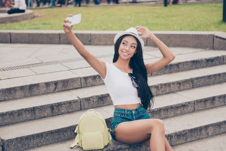 Cute lady with bronze skin and long dark hair, in cap is making a selfie shot while outside in town. She sits on concrete rung, holds headwear, in short jeans denim shorts, in white topの写真素材