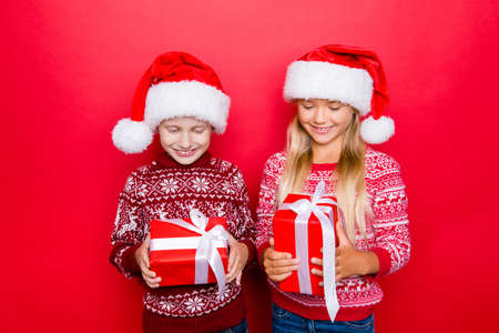 Small adorable charming relatives in traditional x mas clothing, isolated on red background, excited, looking at gifts, hold them and guess what`s inside, curiousity, wish, dream, imagination conceptの写真素材