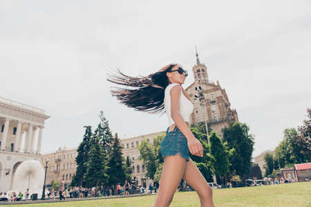 Carefree cute student in summer hot outfit is so happy, jumping outside while on stroll in town, architecture and green trees background, pure clean long healthy black hair flying in the airの写真素材