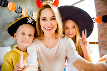 Cropped close up family portrait of three at halloween party. Happy blonde mommy and her cheerful kids - blond small witch and pirate, bonding, lady is making selfie, siblings showing like signsの写真素材