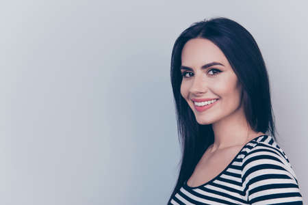 Close up cropped portrait of young charming lady who looks in camera and grinning, on light grey background, in striped fashionable trendy wear, with straight silky dark hair, smooth clean skinの写真素材