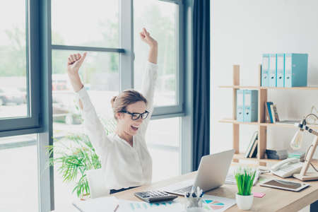 Deal, income, profit, feminity, triumph, victory, happiness concept. A dream of cheerful excited stylish lady leader came true! she is sitting at her light modern work station, celebratingの写真素材