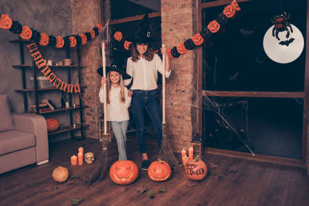 Attractive enchantress mum in elegant clothing, long worlock headwear, smart formal wear and jeans, and charming blonde girl, having a broom sticks in arms, in room with garlands, loft styleの写真素材