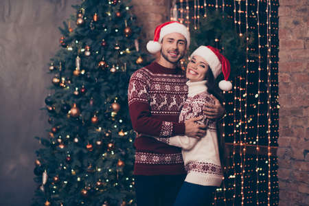 Adorable sweet caucasian husband and wife are bonding, so excited in knitted cute traditional x mas costumes with ornament, headwear, jeans, cuddle, enjoy, garland of sparkles on window, decemberの写真素材