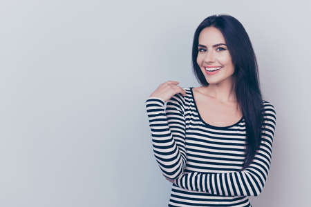 Close up portrait of young charming lady who looks in camera and smiling, on light grey background, in striped fashionable trendy wear, with long silky dark hair, smooth clean skinの写真素材