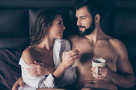 Enjoy each other, mug of tea. Two lovers are cuddling in the bed, looking deep into eyes of each other, true love, feelings, emotions, he is virile mature bearded, she is cute with curly hairの写真素材