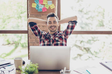 Cheerful young brunet guy is resting in front of his laptop screen, at his work place, with arms behind the head, smiling, with closed eyes, serene and peaceful break in the officeの写真素材