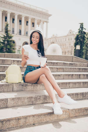Attractive carefree gorgeous mixed-race lady with bronze skin is on stroll, in jeans short shorts, so fit and slim body, chilling with hot tea, on concrete rung, writing message on her pda, sunny dayの写真素材