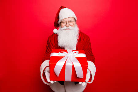 Holly jolly x mas! Festive seosonal occasion. Funny Saint Nicholas in red traditional fur coat presenting a wrapped gift with white tape bow, isolated on red background, wishing happinessの写真素材
