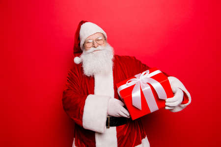 Happiness, noel, festive seosonal occasion. Funny Saint Nicholas in red traditional fur coat and head wear with a wrapped gift with bow, isolated on red background, wishing holly jolly x masの写真素材