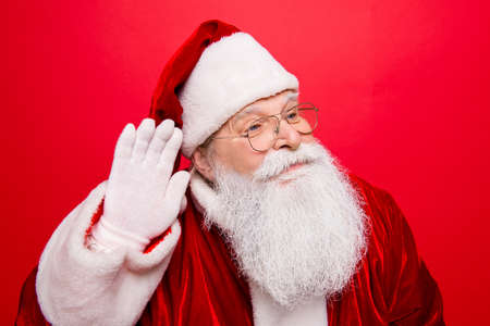 Holly jolly x mas, noel! Festive seosonal time. Funny cheerful saint nicholas in traditional head wear shows hand on ear gesturing with arm, gifts and presents list, isolated on red backgroundの写真素材