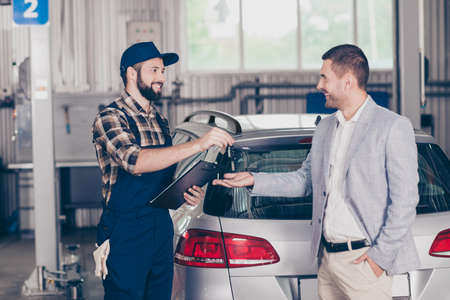 Side profile shot of cheerful professional repairman in cap and uniform, presenting keys of silver car to classy man owner. Car breakdown, check condition, inspection, engineering, insuranceの写真素材
