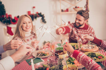 Close up cropped shot of married couple gathered with relatives, with nice setted festive desktop with tasty food plates, treats, dishes, arms putting sticks of sparklers togetherの写真素材