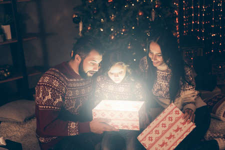 Astonished excited cheerful dreamy girl kid and parents look inside illuminated box, in dark room. Happiness, friendship, wonder, x mas celebrate night, sale promotion, dream giftの写真素材