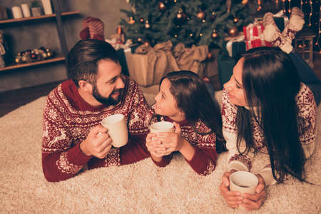 Closeup of three cheerful beautiful relatives at home, chill, relax, celebrate christmastime, girl with mom and dad, in knitted x mas costumes, having cups of  coffee, chocolate, milk to warm-upの写真素材