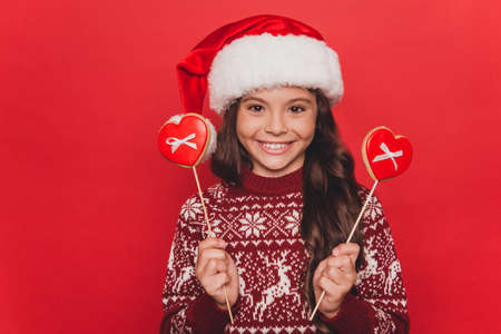 Portrait of pretty small adorable charming girl with cookies in shape of heart, wearing knitted traditional costume, x mas eve celebration, stand isolated on pure red backgroundの写真素材