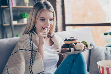 Portrait of beautiful emotional charming attractive sweet toothy woman sitting on sofa in living room, holding plate of donuts and macaroons, looking exciting satisfiedの写真素材