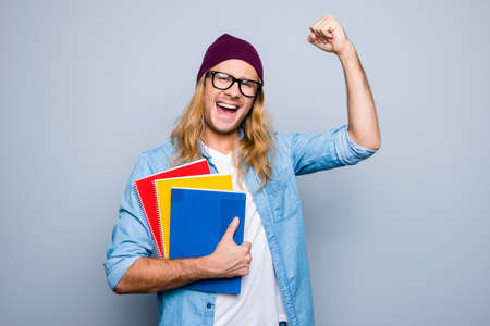 Portrait of glad guy raising his hand with fist having good mood celebrating victory holding three colorful copy books standing over grey background. Victory and triumph conceptの写真素材
