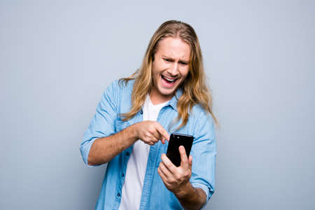 Portrait of angry guy screaming pointing with forefinger to mobilephone standing over grey backgroundの写真素材