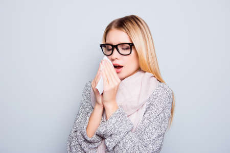 Concept of catching a cold in autumn. Close up portrait of young woman suffering from a cold. She is sneezing and using a paper tissue. She is wearing warm cardigan and scarf. Isolated on backgroundの写真素材