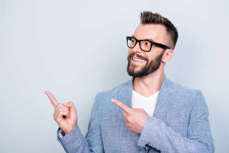 Close up portrait of young successful brunete bearded stock market broker guy on the pure light background, he is smiling, wearing suit and is pointing on a copyspace with his fingersの写真素材