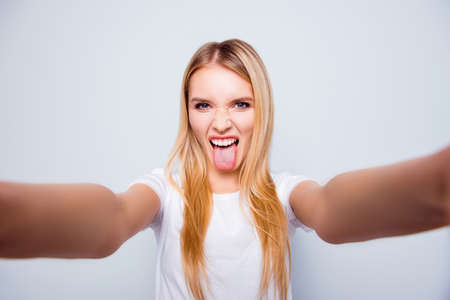 Funky crazy woman with blonde hair is showing her tongue and taking self portrait on her smartphone. She is isolated on grey backgroundの写真素材