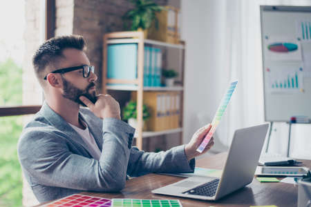 Creative occupation, making decision, looking at the pantone. Side view profile shot of stylish bearded male designer with mustache in trendy specs and grey formal jacket, chooses colors for projectの写真素材