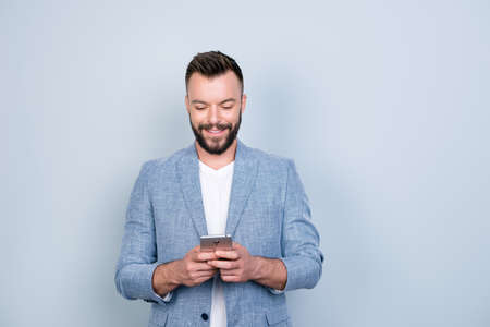 Young cheerful brunete bearded lawyer is standing on the pure light blue background and smiling, reading good news on internet at his mobile phone, wearing casual smartの写真素材