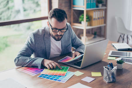 Creative occupation, making decision. Close up of stylish bearded male architect with  mustache in trendy specs and grey formal jacket, choosing the colors for new project, looking at the pantoneの写真素材