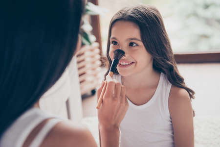 Close up portrait of beautiful smiling little girl with long dark hair, her mother is applying doing make up and applying powder on her nose using a brush, they are having funの写真素材