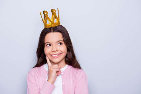 Close up portrait of little caucasian cute girl with crown on head looking up holding forefinger near face dreaming to be a queen while standing over grey backgroundの写真素材
