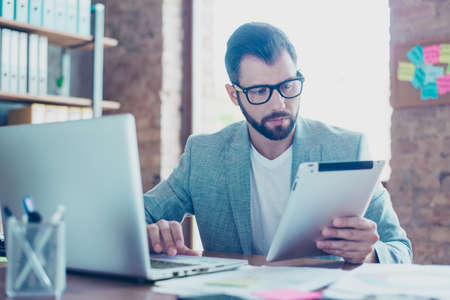 Portrait of young, serious, IT specialist holding tablet in one hand, working through the internet on computer, sitting at his desktopの写真素材