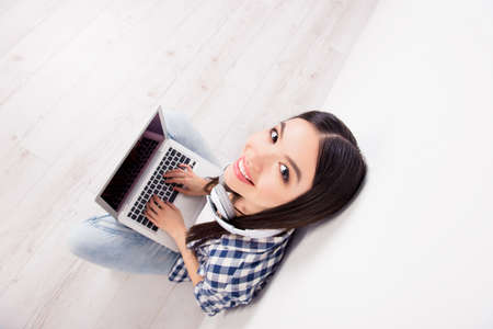 Top view of cute smiling, pretty girl looking to camera, having laptop on her crossed-legs, writing...