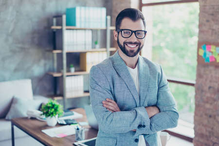Portrait of young, happy economist with beaming smile and stubble after work, standing in workstation, having his hands crossedの写真素材
