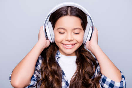 Close up portrait of brunette charming smiling kid holding headphones on her head enjoying her favorite music with close eyes standing over grey backgroundの写真素材