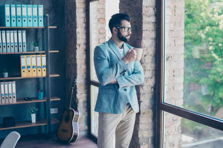 Portrait of young, attractive, serious lawyer, standing with mug of tea near window of his office, looking at window, thinking about workの写真素材