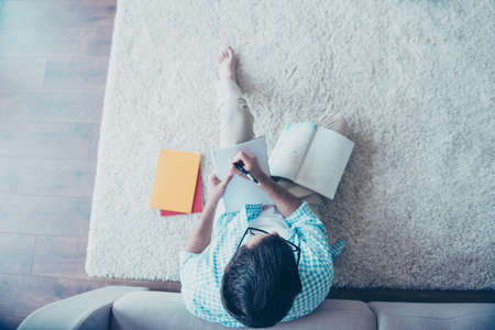 Top view of man in spectacles writing tasks in copybook on his legs, sitting on the floor near couch, preparing for lessonsの写真素材
