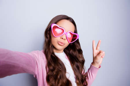 Close up self-portrait of pretty little girl making selfie gesturing peace symbol in casual clothes and  heart-shaped glasses while standing over grey backgroundの写真素材