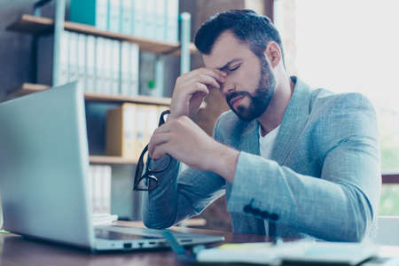 Portrait of attractive young IT person, employee taking off his spectacles, having eyes hurt, having his eyes closed, holding his fingers on nose between eyes, looking tired, overworkedの写真素材
