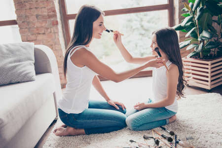 Concept of spending time together with kids! Happy beautiful mother and her little cute daughter are sitting  on the floor in casual clothes, they are holding brushes and doing make up for each otherの写真素材