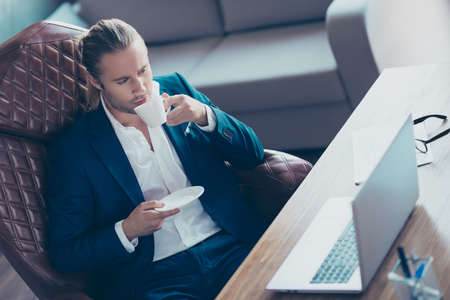 Portrait of attractive manager sitting at his desk in work place, holding mug of tea, drinking...