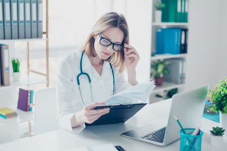 Experienced qualified confident doctor in white coat and spectacles is holding documents and analyzing them, sitting at the table in front of computer, the workplaceの写真素材