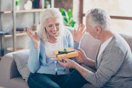 Happy senior couple having anniversary day, bearded husband presenting a packing gift for his wife,...