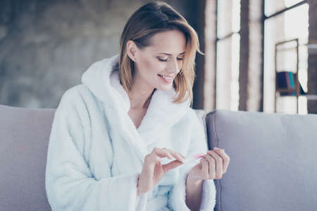 Portrait of cheerful happy smiling woman with short blonde hair dressed in white bathrobe, she is doing manicure and using nail fileの写真素材