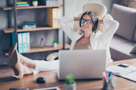 Concept of finishing projects and hard tasks. Pretty beautiful happy satisfied agent in white suit is sitting at the table and relaxing at the workstationの写真素材