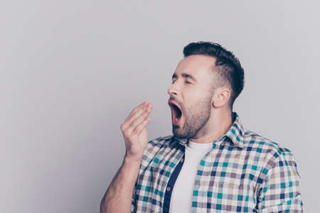 Portrait of tired, bored, attractive man in checkered shirt, closing his open mouth, he wants to sleep, yawning over grey backgroundの写真素材