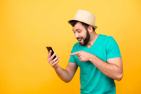 Ha-ha! So funny! Portrait of happy excited crazy man with stubble wearing hat and green tshirt, he is pointing on his smartphone and rejoicingの写真素材