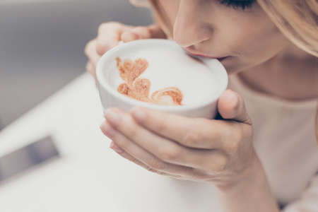 Perfect start of day! Close up white aromatic coffee cup with heart shape latte art foam drinking blonde womanの写真素材