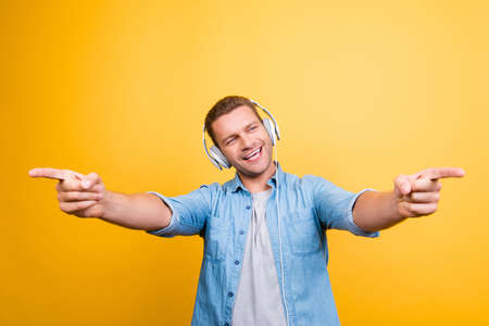 Portrait of caucasion, positive guy in jeans shirt with bristle listening his favorite music, singing the song, pointing index fingers front over yellow backgroundの写真素材