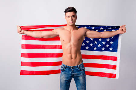 Portrait of confident handsome shirtless guy wearing jeans, he is holding flag of the USA on his back, isolated on grey backgroundの写真素材